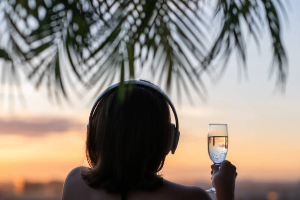 widok z tyłu sylwetka zrelaksowanej kobiety ze szkłem champagn i słuchawkami słuchających muzyki na plaży o zachodzie słońca w gałęziach palm. - tourist resort audio zdjęcia i obrazy z banku zdjęć