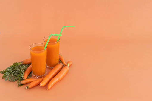 two glasses of carrot juice with a cocktail tube and a lot of carrots on an orange background