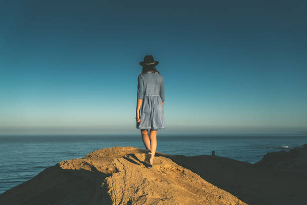 Seascape. Young woman standing on a cliff, calm sea in background. Wearing hat and summer dress. Rear view