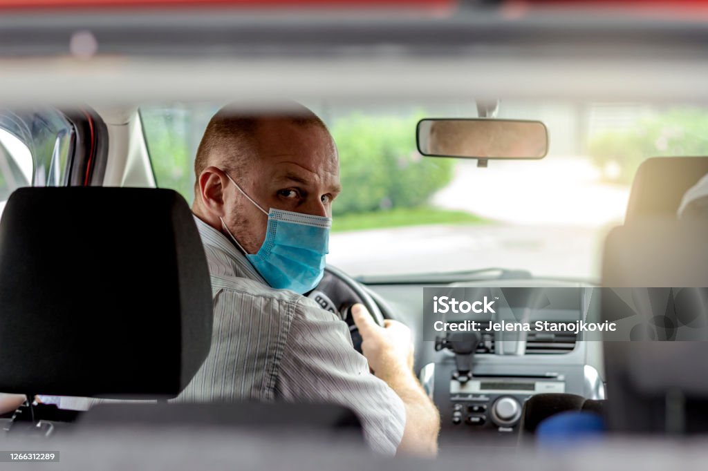 Man with protective mask in a car. Photo of handsome young man wearing face mask sitting in a car,view from rear seat. Driver taking to a passenger on seat back wearing protective medical mask. One man with protective mask riding a car Taxi Driver Stock Photo