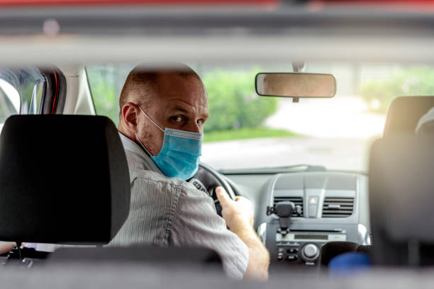 Man with protective mask in a car. Photo of handsome young man wearing face mask sitting in a car,view from rear seat. Driver taking to a passenger on seat back wearing protective medical mask. One man with protective mask riding a car taxi driver photos stock pictures, royalty-free photos & images