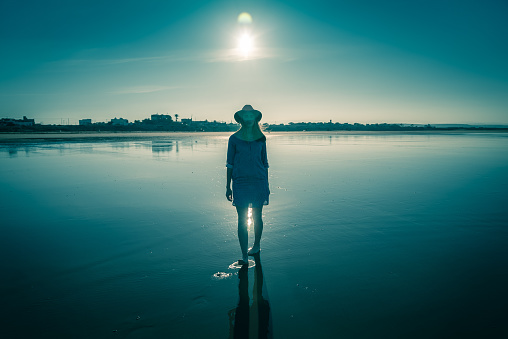 Seascape. Young woman standing on sand, calm sea in background. Wearing hat and summer dress