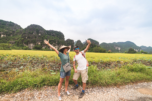 Couple selfie on countryside road among rice fields. Man with backpack and woman with vietnamese hat having fun traveling together on vacation. Tam Coc Trang An Ninh Binh Vietnam.