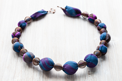 handcrafted necklace of round beads wrapped in blue silk cloth and metal spiral beads on wooden table close up