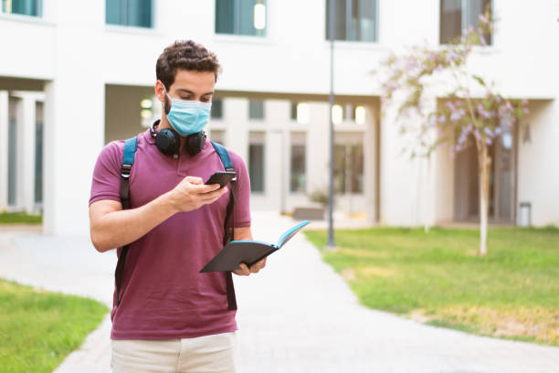 Student wearing a surgical mask taking a photo of his notebook with a mobile phone. Coronavirus pandemic concept. stock photo