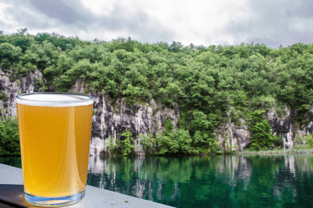 стакан светлого пива с видом на изумрудное озеро в хорватии». nlager пиво против крутых берегов с деревьями. - parks brewery стоковые фото и изображения
