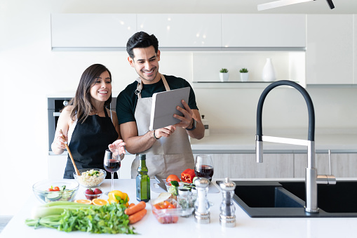 Happy Hispanic couple looking recipe on internet through digital tablet in kitchen