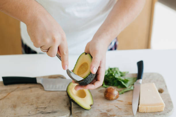 Person peeling perfectly ripe avocado with spoon for sandwich on modern white kitchen. Process of making healthy toasts with avocado, tomato, arugula, cheese. Home cooking concept stock photo