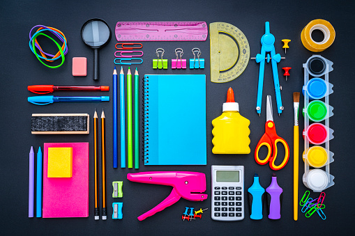 Back to school concepts: overhead view of a large group of multicolored school supplies shot on black background. The composition includes a drawing compass, color pencils, paint brushes, paper clips, pencil sharpener, eraser, crayons, pocket calculator, thumbtack, rubber bands, notepad, stapler, adhesive tape, felt tip pen and scissors. High resolution 42Mp studio digital capture taken with SONY A7rII and Zeiss Batis 40mm F2.0 CF lens