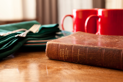 Table is set in cozy home for sharing coffee, a snack and Bible study.   Red coffee mugs, green cloth napkins, forks and a well used Bible.
