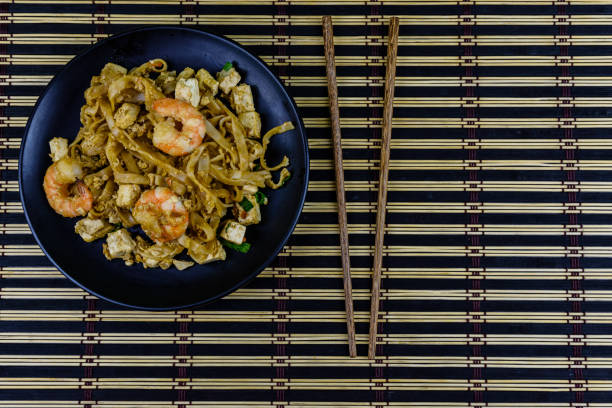 Black plate with pad thai and chopsticks on bamboo mat. Top view