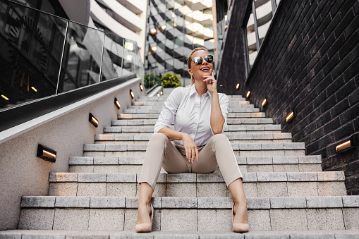 Beautiful smiling fashionable businesswoman sitting on the stairs in front of business center and posing.