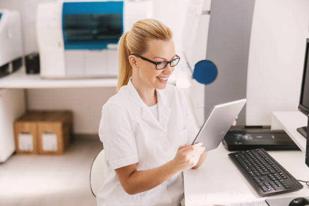 attractive blond lab assistant in sterile uniform with eyeglasses sitting and using tablet for entering test results. - laboratory doctor white collar worker research imagens e fotografias de stock