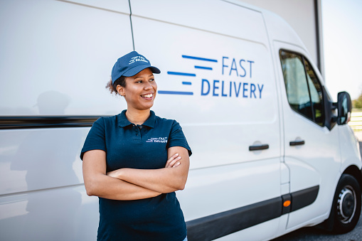 Mixed race female delivery driver in uniform with crossed arms standing next to van and looking away from camera.