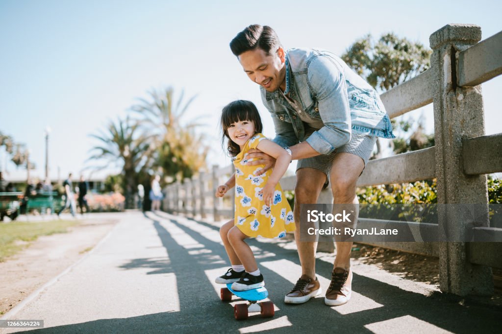 Father Helps Young Daughter Ride Skateboard A dad helps his little girl go skateboarding, holding her waist for support.  Shot in Los Angeles, California by the Santa Monica Pier. Family Stock Photo