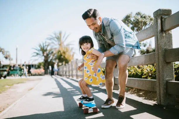 A dad helps his little girl go skateboarding, holding her waist for support.  Shot in Los Angeles, California by the Santa Monica Pier.