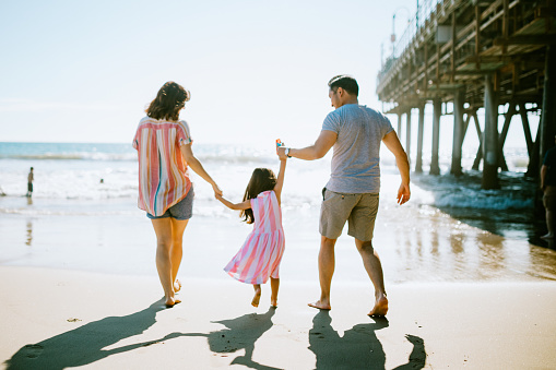 A cute young family walks along the sand by the Santa Monica Pier in Los Angeles, California.  The father and mother hold their daughters hands, lifting her up when the waves come in.  A much needed break from the city in the ocean air.