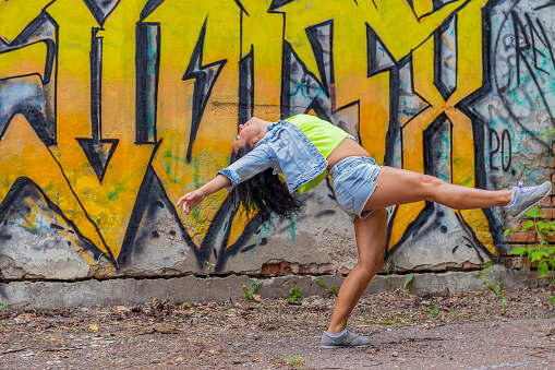 A young woman in a denim suit and a bright T-shirt bent back in a dance against a wall with a yellow graffiti pattern