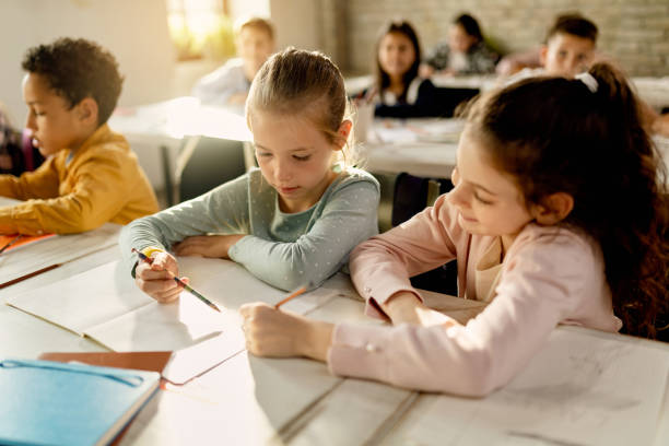 Two schoolgirls working together on a assignment in the classroom. Female classmates learning together at their desk during a class at elementary school. elementary school building photos stock pictures, royalty-free photos & images