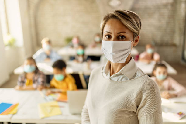 Elementary school teacher with protective face mask in the classroom. Female teacher wearing a face mask while teaching children at elementary school and looking at camera. social responsibility photos stock pictures, royalty-free photos & images
