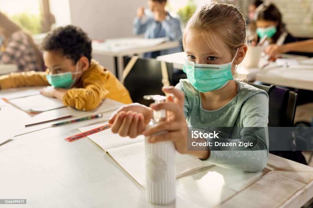 Elementary student wearing protective face mask and disinfecting her hands in the classroom. Schoolgirl with face mask using hands sanitizer while sitting at a desk in the classroom. Child Stock Photo