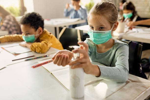 Elementary student wearing protective face mask and disinfecting her hands in the classroom. Schoolgirl with face mask using hands sanitizer while sitting at a desk in the classroom. reopening photos stock pictures, royalty-free photos & images