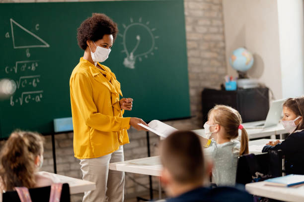 Happy black teacher giving exam paper to her student while wearing protective face mask in the classroom. Happy black teacher and her students wearing protective face mask in the classroom. Teacher is giving them their test results. person wearing n95 mask stock pictures, royalty-free photos & images