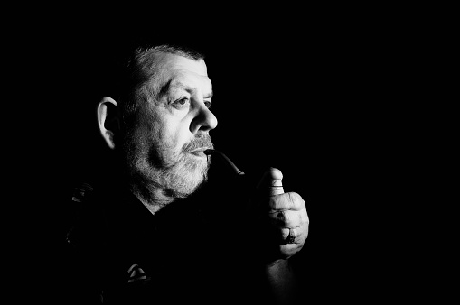 Nice Black and white dramatic portrait of caucasian senior smoking tobacco pipe in darkness