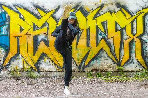 Young girl in stylish black overalls makes a high kick against the background of a concrete wall with a yellow graffiti pattern