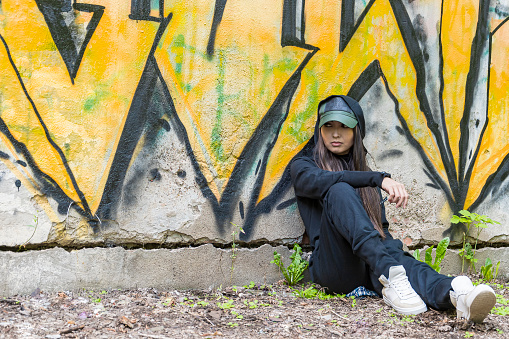 A young girl in a stylish black jumpsuit sits and rests against a concrete wall with a yellow graffiti pattern