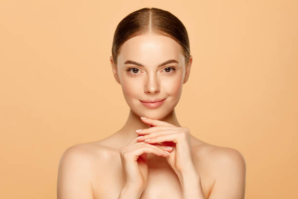 young beautiful smiling female standing with naked glowing skin and shoulders, touching chin, isolated on beige background. woman beauty concept - clear sky human skin women smiling imagens e fotografias de stock