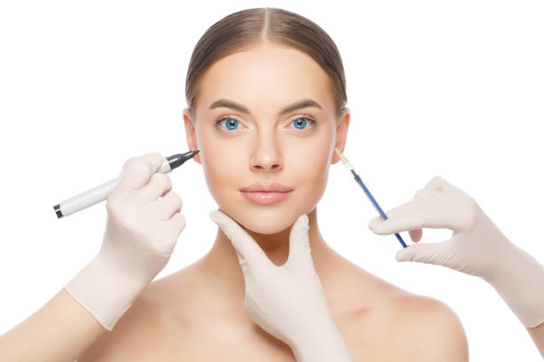 istock Young blue-eyed female ready for beauty procedures from cosmetologists whose hands in gloves hold skin marker and injection syringe, isolated on white background 1266420176