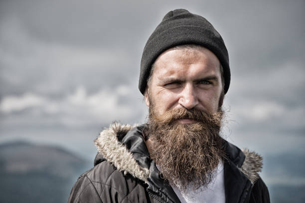 istock Man with long beard and mustache wears hat. Hipster on strict face with beard looks brutally while hiking. Masculinity concept. Man with brutal bearded appearance, brutal unshaven man looks untidy 1266422597