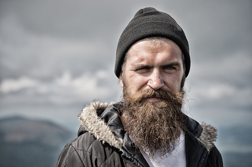 Man with long beard and mustache wears hat. Hipster on strict face with beard looks brutally while hiking. Masculinity concept. Man with brutal bearded appearance, brutal unshaven man looks untidy