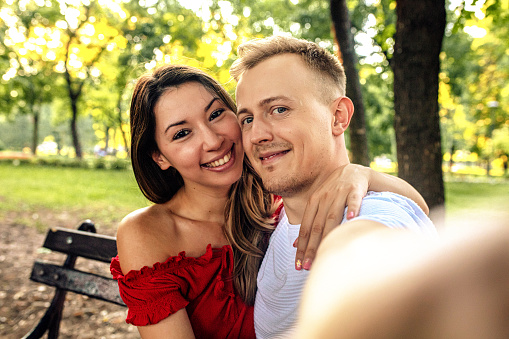 Cheerful young couple taking selfie sitting on bench outdoors