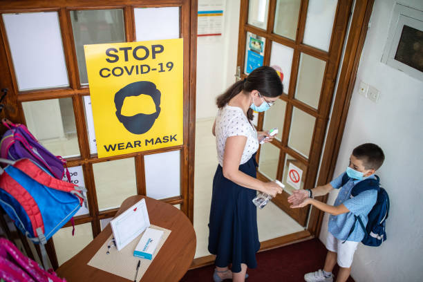 Teacher spraying antiseptic on schoolboy's hands before he enters school during coronavirus pandemic Teacher spraying antiseptic on schoolboy's hands before he enters school, they are wearing protective face masks for protection against virus during covid-19 pandemic
, poster about virus prevention is on doors backpack sprayer stock pictures, royalty-free photos & images