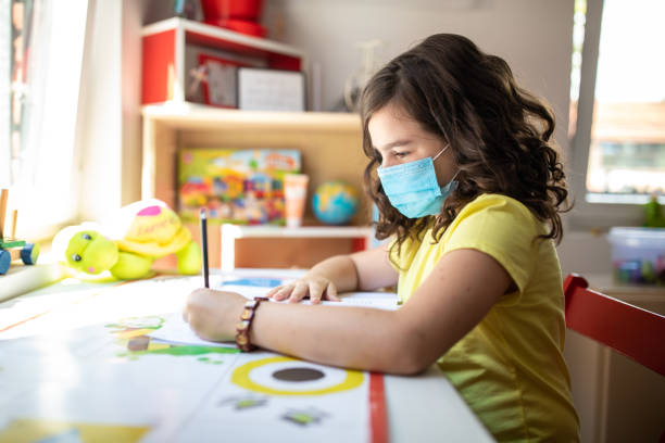 istock Schoolgirl with protective face mask sitting at desk and writing in note pad 1266458699