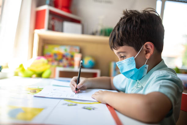 istock Schoolboy with protective face mask sitting at desk and writing in notepad 1266458709
