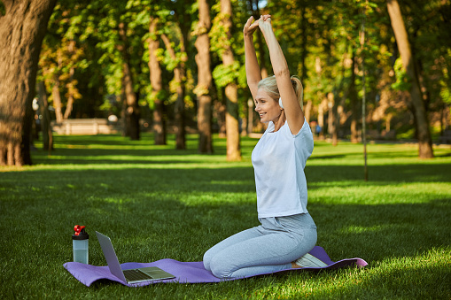 Cheerful sporty lady doing exercise and smiling while sitting on yoga mat in front of notebook