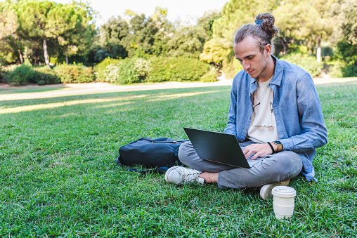 trendy caucasian man sitting on the grass and using a modern laptop