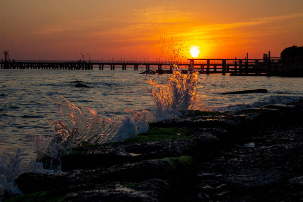 Beautiful splashes of water breaking on concrete slabs, breakwaters, waves. The water is illuminated by the outgoing sun. Bright orange sunset on the sea. There's a pier in the distance.
