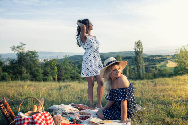 Two beautiful women are enjoying the summer time, organizing a picnic with food, wine and a beautiful view of the nature