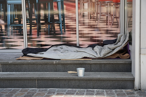 Bolsa de dormir de una persona sin hogar esparcida en las escaleras frente a un moderno salón o restaurante elegante. Hay una pequeña copa de metal cerca de la litera pidiendo limosna y caridad. photo
