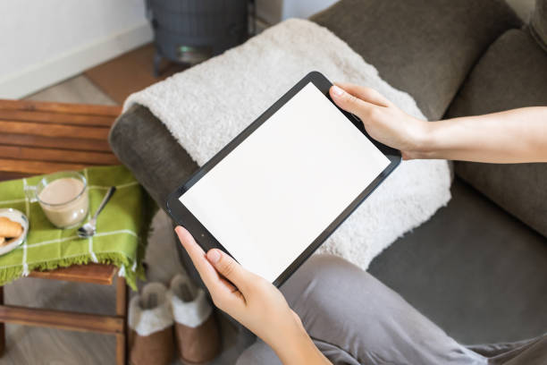 Hands hold a black digital tablet on the living room sofa. Tablet mockup with blank space for advertising