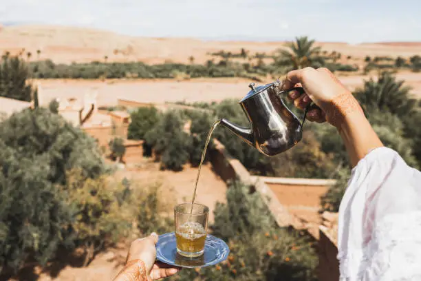 Photo of Woman hands pouring glass of traditional mint moroccan tea with vintage silver tableware. Sand desert and palms on background.