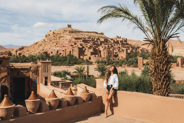 beautiful young woman happy to travel in morocco. ait-ben-haddou kasbah on background. wearing in white shirt and jeans shorts. - journey elegance people traveling architecture stock-fotos und bilder