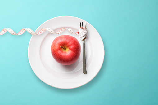 Red Apple on a plate and a tape measure rolled up a fork on light blue background.\nDiet concepts.