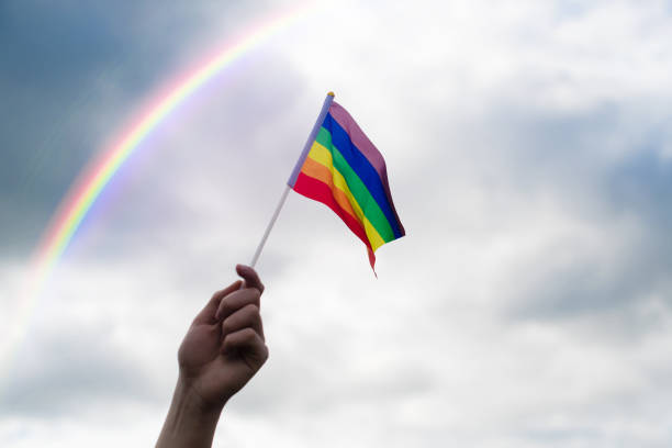 A hand holds an LGBT flag against a sky with clouds and a rainbow. stock photo