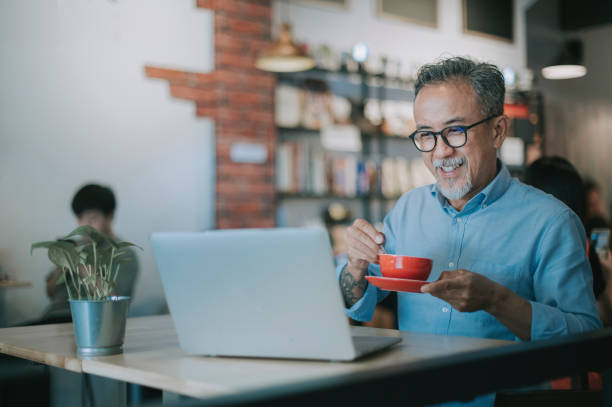 an asian chinese senior man having discussion with his colleague via online virtual meeting in a cafe an asian chinese senior man having discussion with his colleague via online virtual meeting malaysia office workers stock pictures, royalty-free photos & images