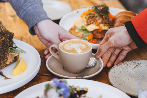 a cup of flat white coffee in cream ceramic cup with latte art on top, touched by two hands, surrounded with variety of food on wooden table stock photo